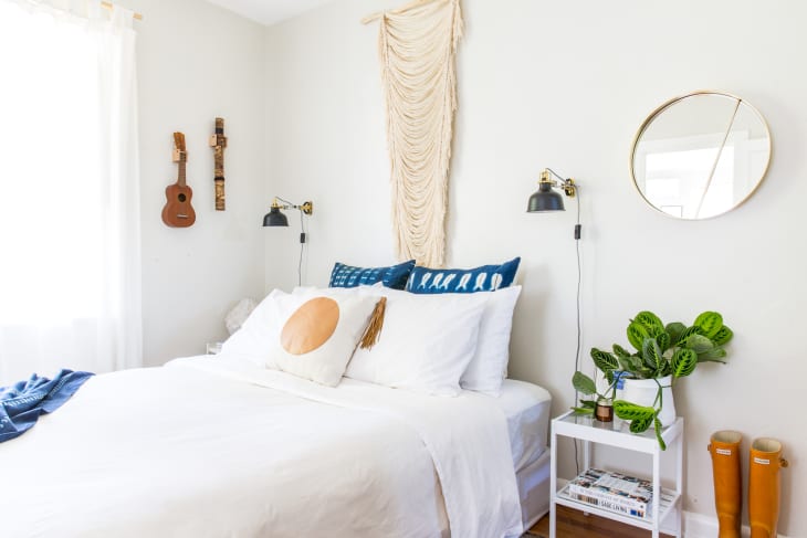 Bedroom with white bedding, blue pillows, wall art, ukuleles, round mirror, and a plant on a bedside table.