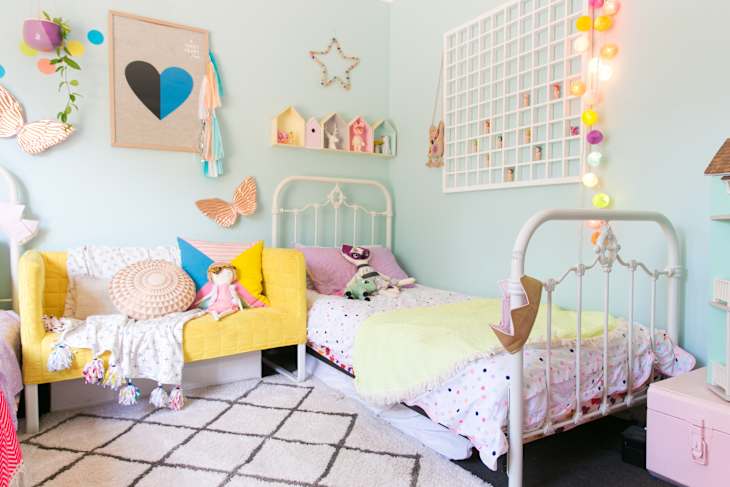 Children's bedroom with pastel decor, twin beds, yellow sofa, colorful pillows, and string lights on a mint green wall.