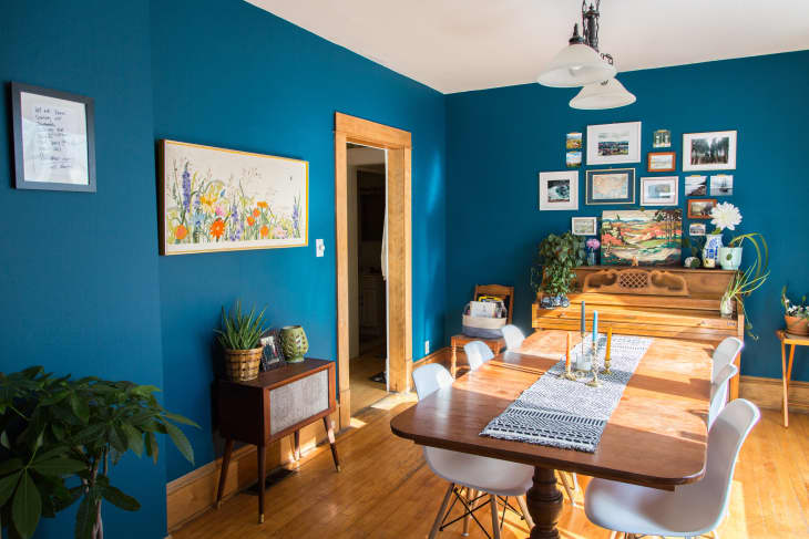 Dining room with blue walls, wooden table, white chairs, wall art, and a piano adorned with plants and decor.