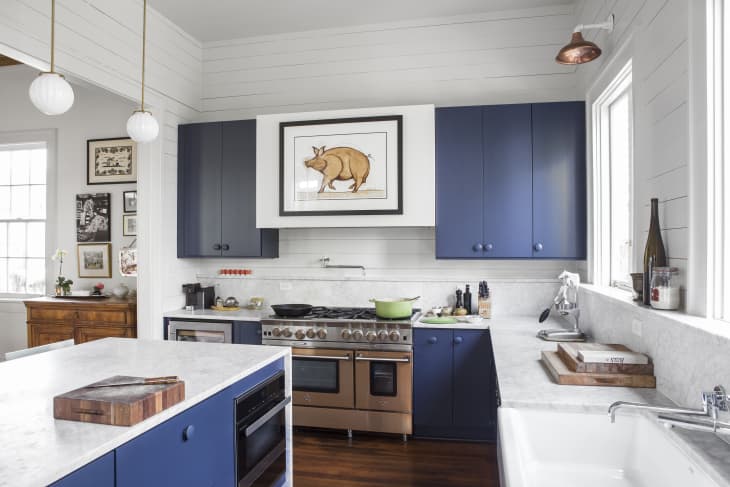 Modern kitchen with blue cabinets, marble countertops, a large stove, and framed pig artwork above.