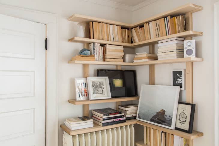 Corner bookshelf with stacked books, framed art, a small speaker, and decorative items on light wood shelves.