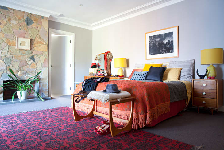 Bedroom with stone fireplace, red patterned rug, orange bedspread, yellow lamps, and wooden side tables.