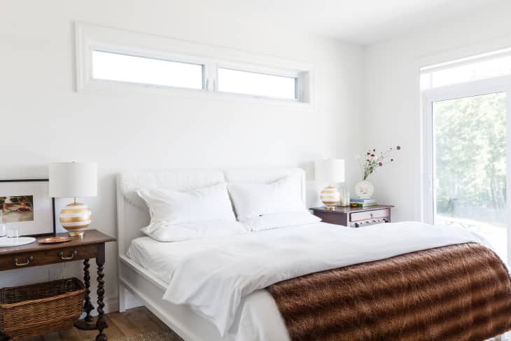 Bright bedroom with white bedding, brown fur throw, wooden nightstands, striped lamps, and a window view of greenery.