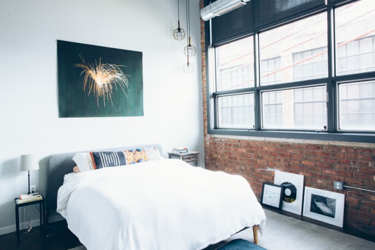 Loft bedroom with a large window, brick wall, modern art, and a bed with a white comforter.