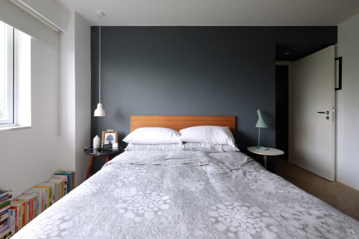 Bedroom with a wooden headboard, gray accent wall, white bedding, bedside tables, and books stacked by the window.