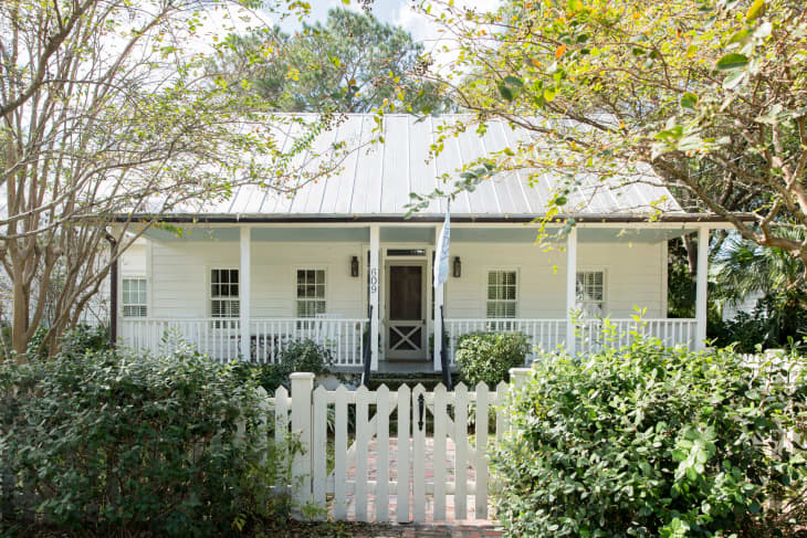 White cottage with a metal roof, front porch, and picket fence, surrounded by trees and shrubs.
