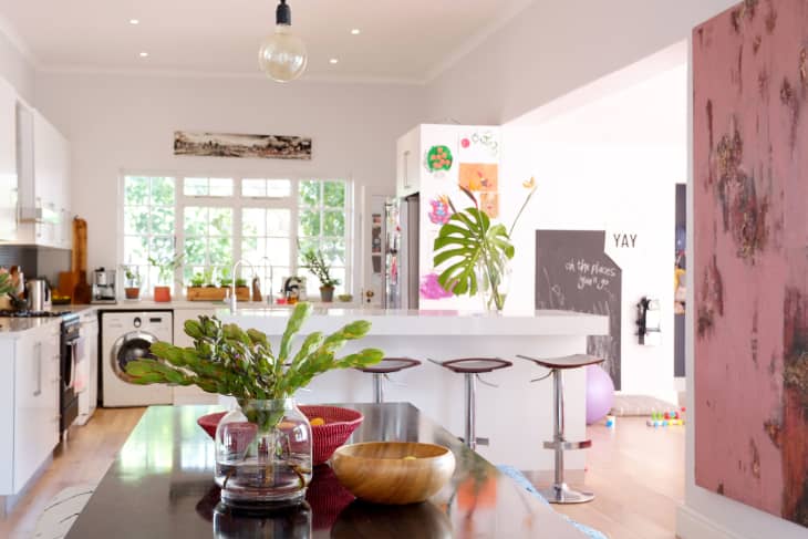 Bright kitchen with white cabinets, island with bar stools, large plant, and colorful artwork on the walls.