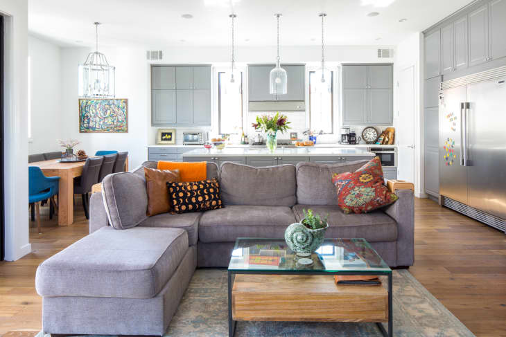 Gray sectional sofa with colorful pillows in open-plan kitchen and dining area, featuring gray cabinets and a glass coffee table.