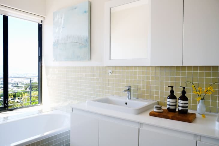 Modern bathroom with white cabinets, a sink, yellow tiled backsplash, and a window view. Soap dispensers and flowers on counter.