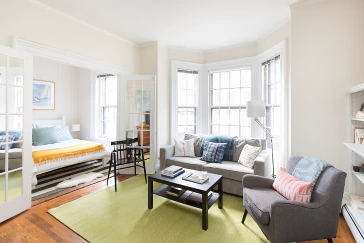 Living room with gray sofa, colorful pillows, armchair, coffee table, and view into bedroom with yellow blanket.