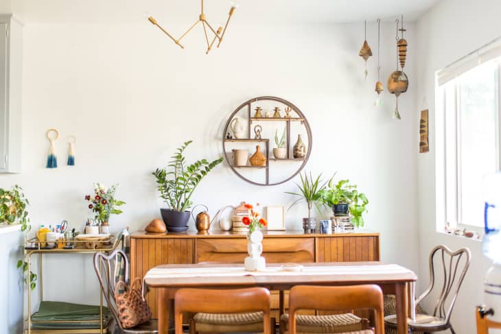 Dining room with wooden table, chairs, plants, and wall decor, including a round shelf with pottery and hanging ornaments.