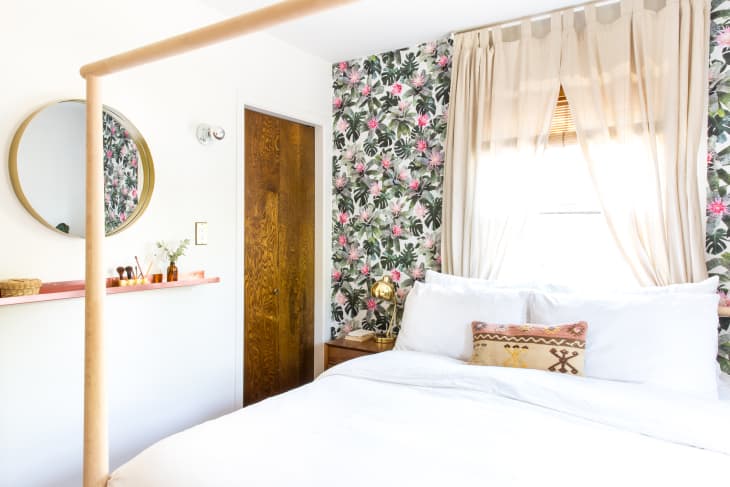 Bedroom with floral wallpaper, white bedding, round mirror, and wooden door. Beige curtains and decorative pillow on bed.
