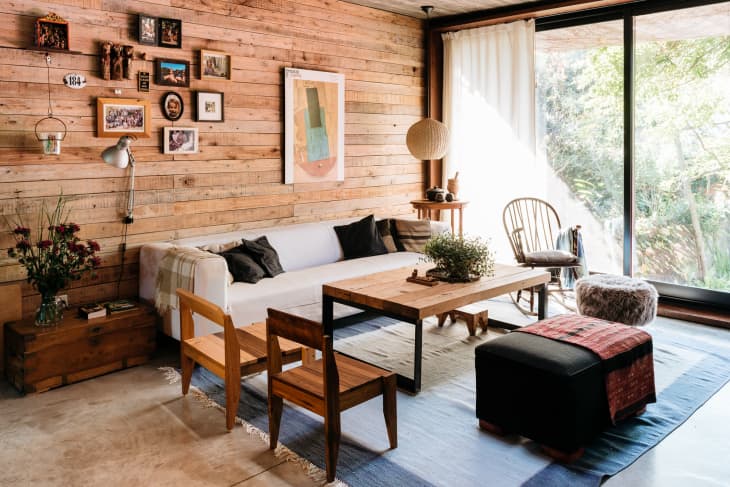 Cozy living room with wooden walls, white sofa, wooden chairs, coffee table, and gallery wall, overlooking a garden through glass doors.