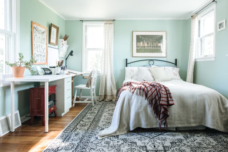 Cozy bedroom with a white desk, potted plant, and a bed with a striped throw blanket on a patterned rug.