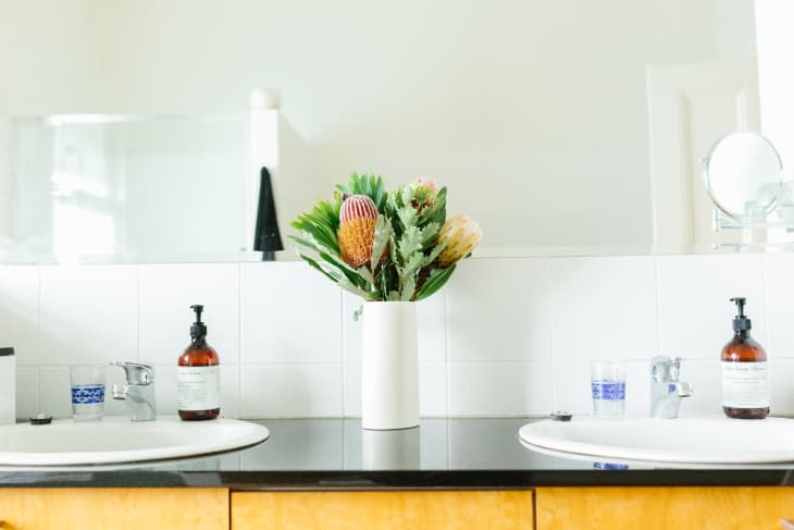 Bathroom vanity with dual sinks, a white vase of flowers, and soap dispensers on a black countertop.