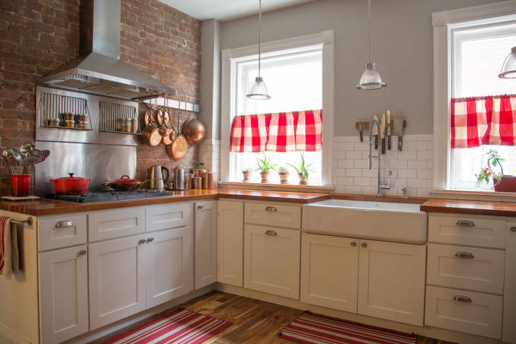 Red buffalo plaid cafe curtains in a white farmhouse kitchen with an apron sink, a stainless steel stove and range hood, an exposed brick wall, and copper pots and pans
