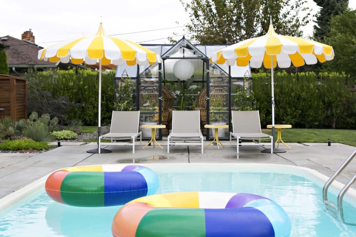 Poolside with colorful inflatable rings, yellow-striped umbrellas, and lounge chairs near a glass greenhouse.