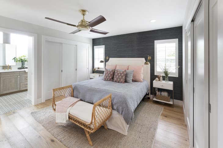 Modern bedroom with gray bedding, pink pillows, wicker bench, and dark accent wall. Ensuite bathroom visible on the left.