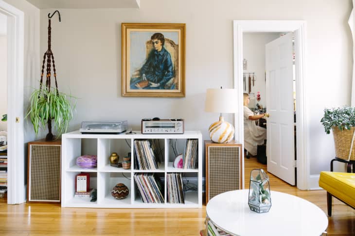 Living room with a white shelf holding records, a turntable, and decor. A painting hangs above, and a person is visible through a doorway.
