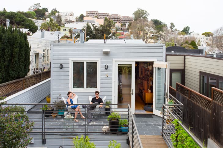 Two people sitting on a rooftop patio with potted plants, outside a small gray house with open French doors.