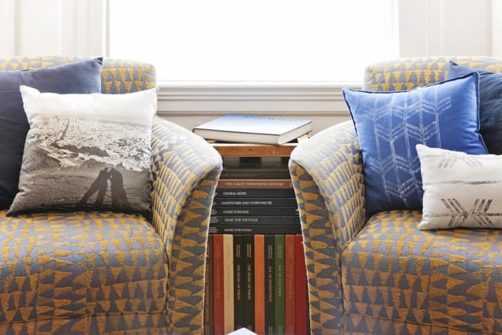 Two patterned armchairs with decorative pillows flank a small table stacked with books.