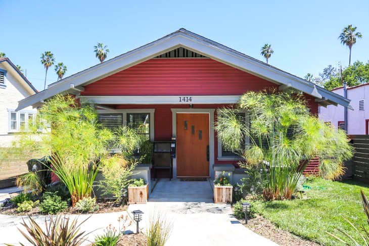 Red bungalow with a wooden door, surrounded by lush plants and a small front yard.
