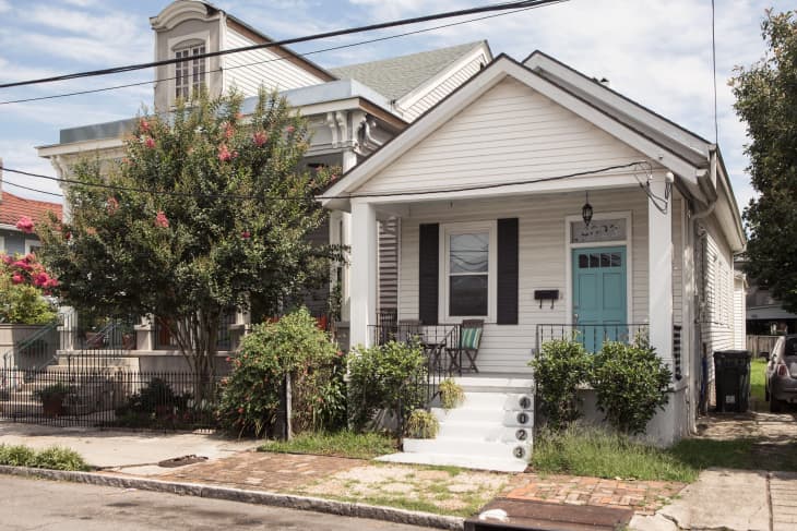 White house with a teal door, black shutters, and a small porch with chairs, next to a flowering tree.