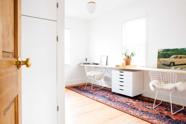 Home office with white desk, wire chairs, fluffy cushions, potted plant, and vintage car poster on a patterned rug.