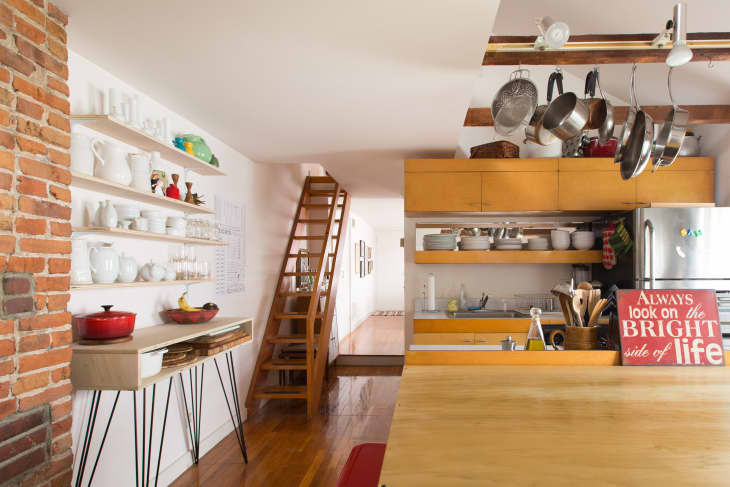 Open kitchen with wooden cabinets, hanging pots, brick wall, and shelves displaying white ceramics and colorful decor.