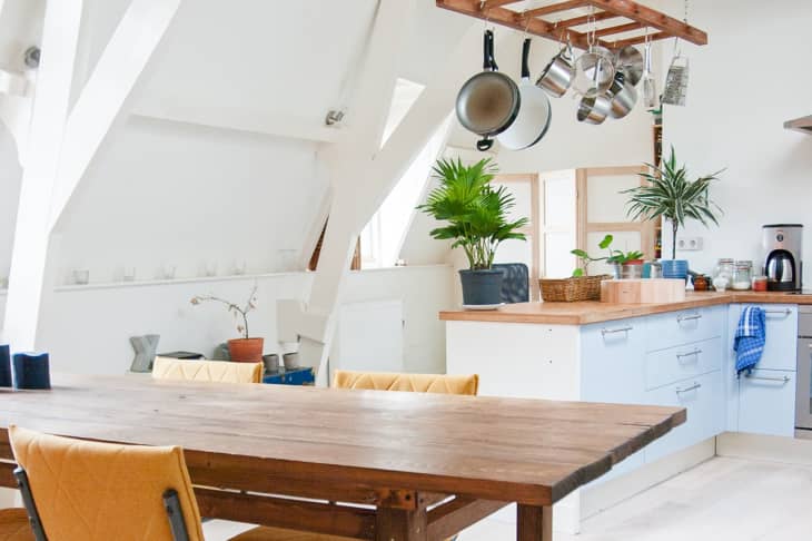 Open kitchen with wooden dining table, hanging pots, potted plants, and light blue cabinets.