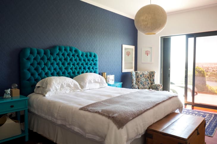 Bedroom with teal tufted headboard, blue accent wall, patterned armchair, and sliding glass doors to a balcony.