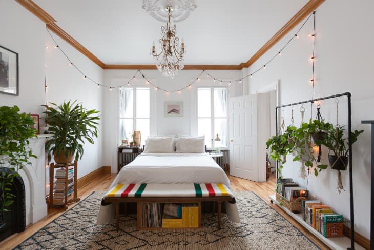 Bedroom with a white bed, hanging plants, string lights, and a colorful striped bench.