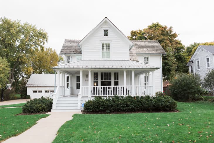 White two-story house with a wraparound porch, surrounded by green lawn and trees.