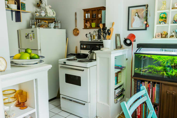 Cozy kitchen with vintage stove, fridge, and a fish tank. Shelves hold books, utensils, and decor. Bowl of citrus on counter.