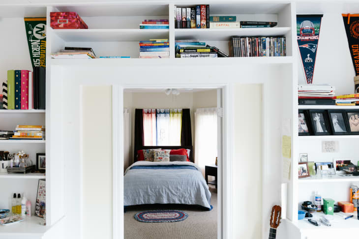 Bedroom with a blue bedspread, colorful curtains, surrounded by bookshelves with books, photos, and sports pennants.