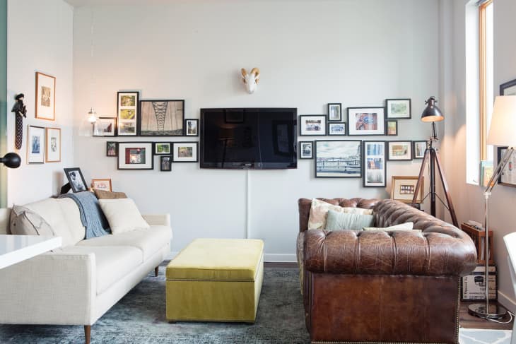 Living room with a beige sofa, brown leather couch, yellow ottoman, wall-mounted TV, and a gallery wall of framed art.