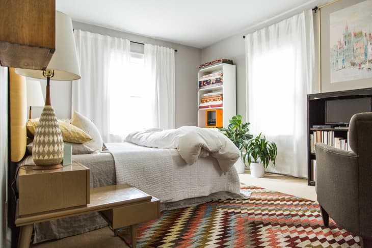 Cozy bedroom with a patterned rug, white bedding, a potted plant, and a bookshelf.