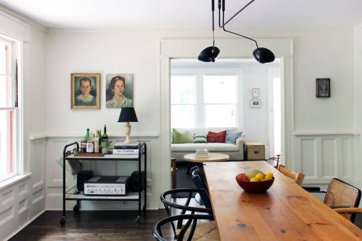 Dining room with wooden table, black chairs, bar cart, and two portraits. Sofa with colorful pillows in the background.