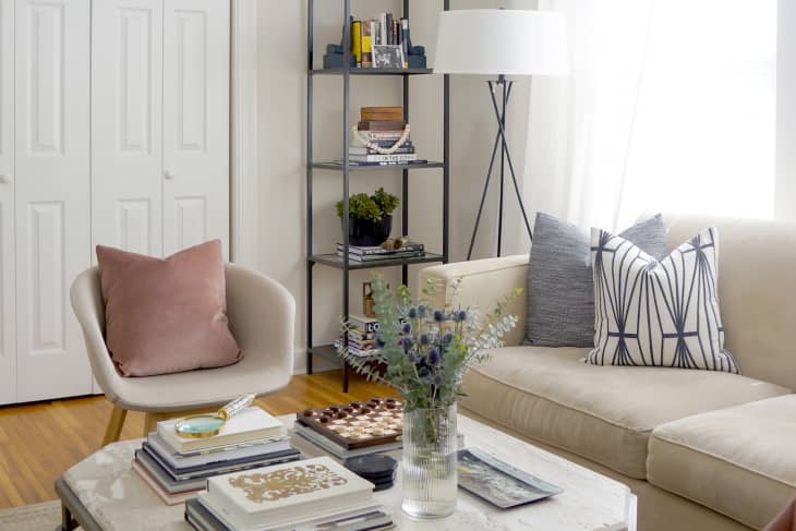 Living room with beige sofa, patterned pillows, pink chair, bookshelves, and a coffee table with flowers and books.