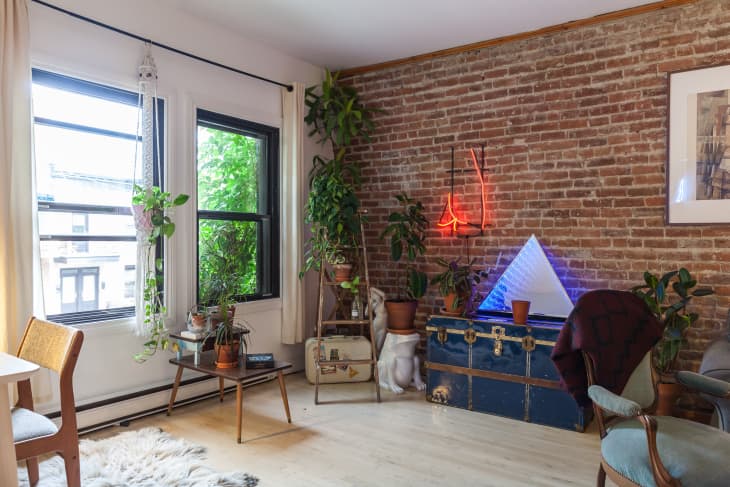 Cozy living room with brick wall, neon lights, plants, vintage trunk, and a triangular light sculpture.