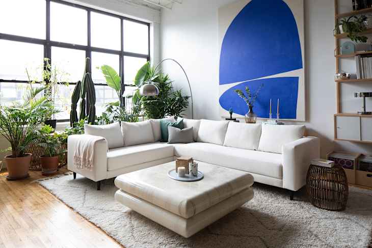 Bright living room featuring a white sectional Sabai sofa, large plants, a blue abstract painting, and a round coffee table.