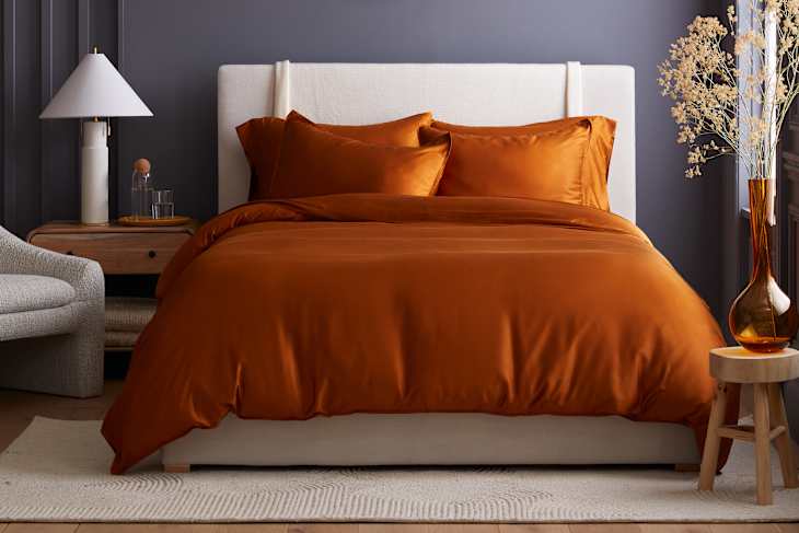 bedroom featuring Quince products: an orange duvet set, white upholstered headboard, and a side table with a lamp and decorative vase.