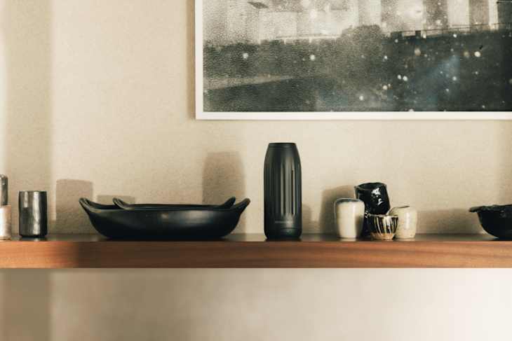 A wooden shelf displaying a black bowl, a textured black vase, and various decorative ceramics against a neutral wall.