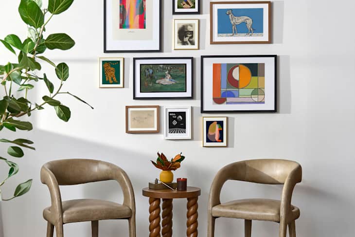 Two beige chairs beside a twisted wooden side table with a small plant and candle, against a gallery wall of framed art.