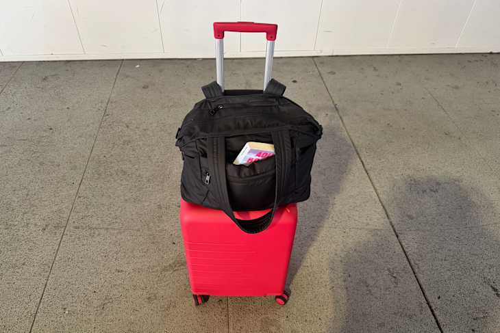 A red suitcase with wheels topped by a black duffel bag, partially revealing a book inside, on a concrete surface.