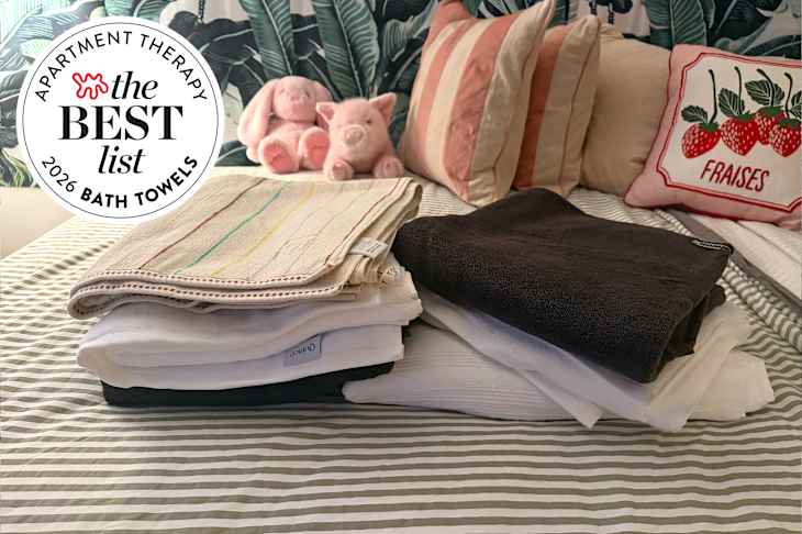 Folded bath towels in various colors on a striped bedspread, with plush toys and decorative pillows in the background.