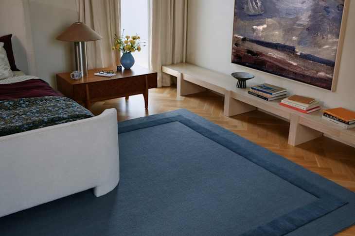 Cozy bedroom featuring a white bed, blue area rug, wooden side table with flowers, and a modern art piece on the wall.