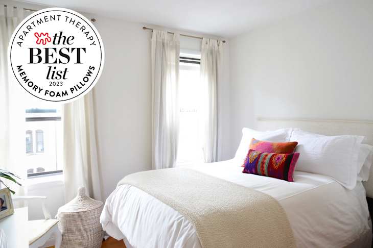 Bedroom with white walls, white curtains. Bed with white bedding and colorful accent pillows. Seal in upper left corner reads "Apartment Therapy The Best List 2023 Memory Foam Pillows"