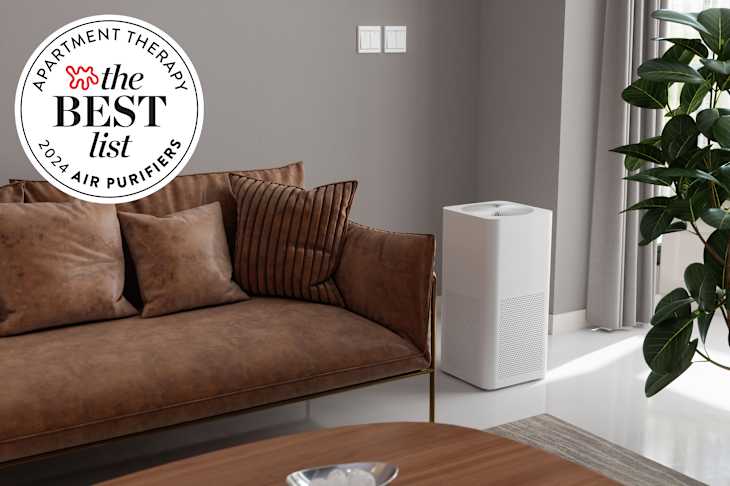 Living room with gray walls, brown leather sofa, potted tree, white floor, wood coffee table, and white air purifier on floor next to sofa. Seal in upper left corner reads "Apartment Therapy The Best List: 2023 Air Purifiers"