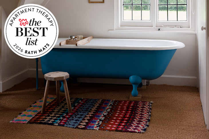 A blue freestanding bathtub with a wooden tray, a small wooden stool, and a colorful patterned rug on a woven floor.